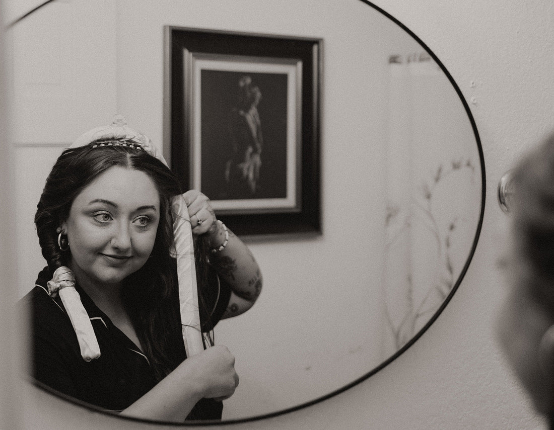 Woman adjusting satin heatless curlers in front of a mirror, demonstrating a simple nighttime no-heat curling routine in a relaxed home setting.