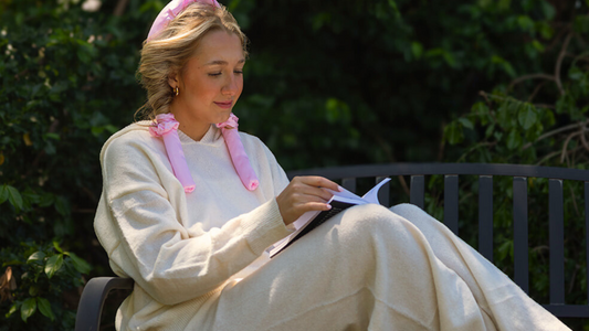 Woman reading on a bench while wearing pink satin heatless curlers, demonstrating effortless no-heat hair styling in a relaxed outdoor routine.
