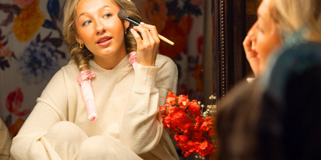 Woman wearing pink satin heatless curlers while applying makeup in the mirror, highlighting multitasking beauty with damage-free overnight curls.
