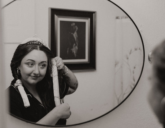 Woman adjusting satin heatless curlers in front of a mirror, demonstrating a simple nighttime no-heat curling routine in a relaxed home setting.